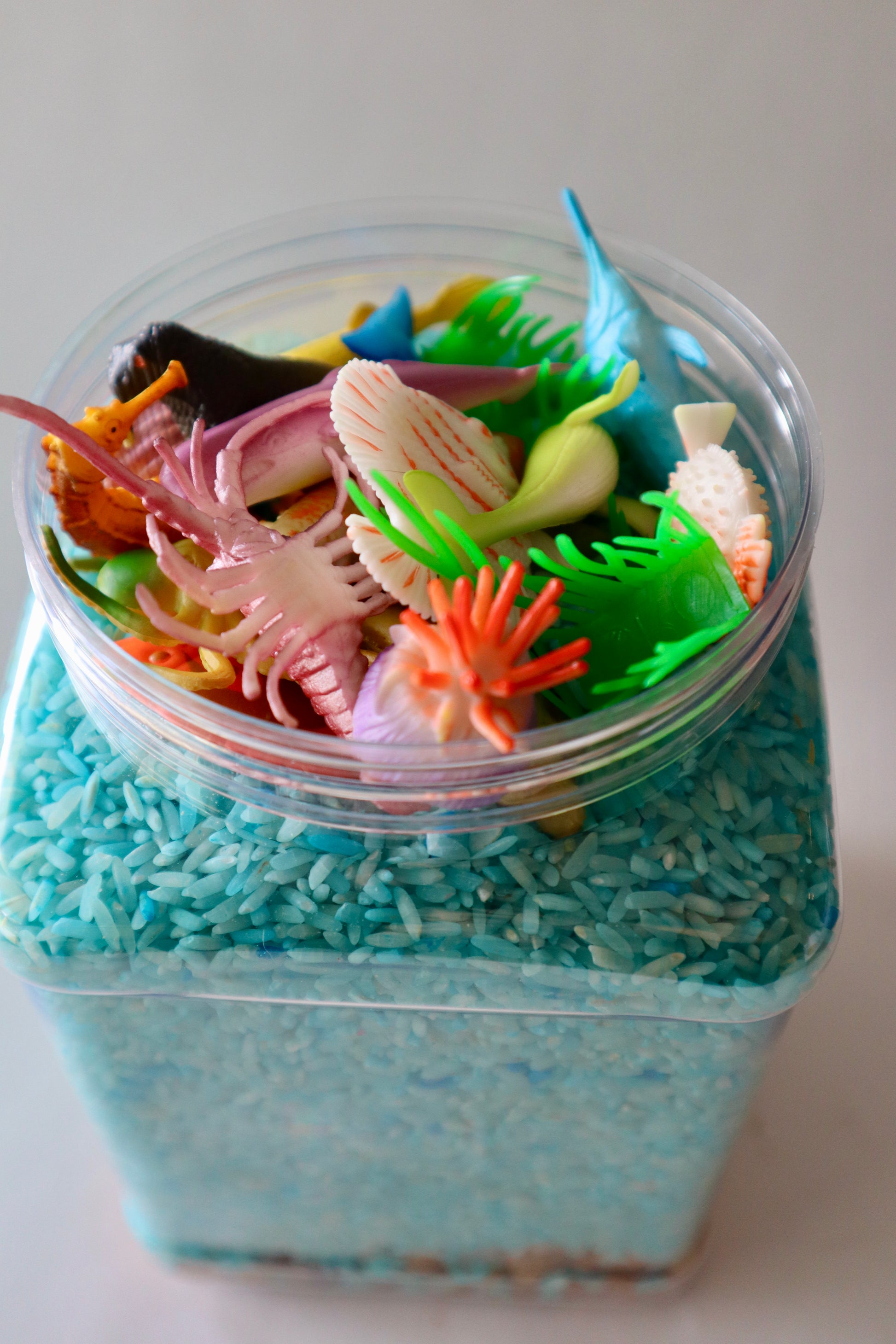 Container filled with colorful miniature sea creatures and blue sand-like material on a white background