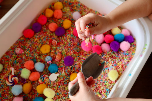 Colorful sensory bin with pom-poms and a child's hand holding a small tool.